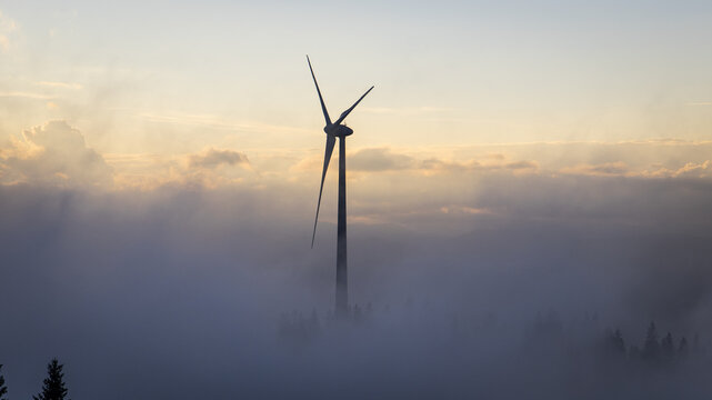 Aerial view of a solitary wind turbine piercing through a sea of ethereal fog, crowned by the warm glow of the setting sun, Gaberl, Styria, Austria.