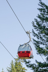 A vibrant red and silver gondola lift cabin is suspended against a clear blue sky, with evergreen trees visible. This iconic transport system is a common sight in the Bansko ski resort. © VikSid