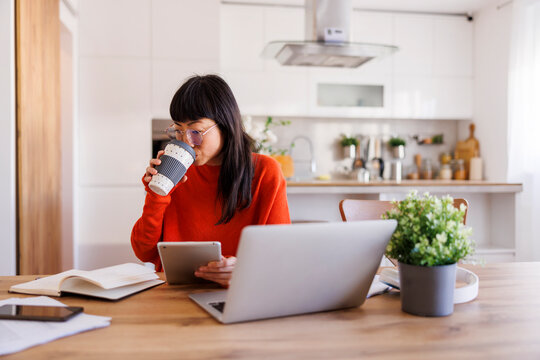 Woman drinking coffee and using tablet and laptop computers while working remotely from home