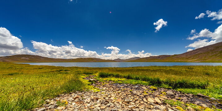 View of a serene lake mirroring the azure sky dotted with fluffy white clouds, bordered by vibrant green meadows and rocky terrain, Deosai National Park, Gilgit Baltistan, Pakistan.