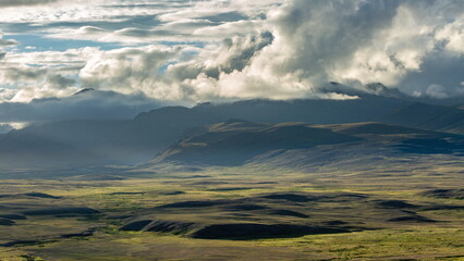 View of a vast, undulating landscape bathed in diffused sunlight, with dramatic clouds casting shadows over distant mountain peaks, Deosai National Park, Gilgit Baltistan, Pakistan.