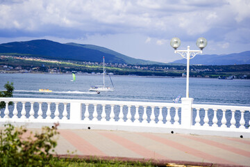 Obraz premium The embankment of the Gelendzhik seaside resort: white balustrade, sea, mountain range in the background. Summer, white clouds
