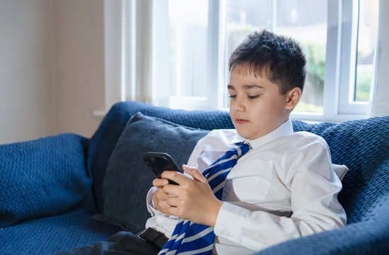 Schoolboy in uniform using a smartphone text message or play game, sitting on blue sofa at home.Back to school Concept of digital learning, technology in education,screen time,after school relaxation - Powered by Adobe
