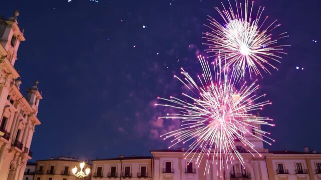 Fireworks illuminate the night sky as fireworks burst over the city, casting a glow on buildings. Buildings and streetlights contrast with fireworks creating a vibrant skyline.