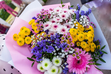 various Flowers bouquet, large petals pink gerbera, chrysanthemum lush yellow, white small aster, kermek dark blue shallow, abundantly blooming, inflorescence, in gift packaging, woman holds in hands,