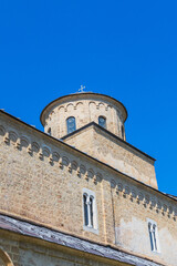 Medieval stone monastery dome with cross, arched windows, under a vibrant blue sky, showcasing Byzantine Orthodox Christian heritage and tranquil Eastern European rural landscape. Studenica, Serbia.