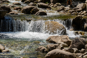 A serene mountain stream in Bansko forest. Clear water flows over rocks, creating small waterfalls and ripples. Sunlight reflects, highlighting its natural beauty.