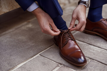 Close-up of a well-dressed man tying a polished brown leather dress shoe while seated on a bench, highlighting hands, laces, classic footwear and tailored navy suit details.