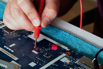 Young computer repairman, laptop motherboard repairman is using IC meter to find faults on motherboard for repair on his desk. Circuit board repair.