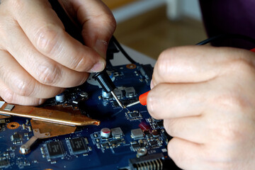 Young computer repairman, laptop motherboard repairman is using IC meter to find faults on motherboard for repair on his desk. Circuit board repair.