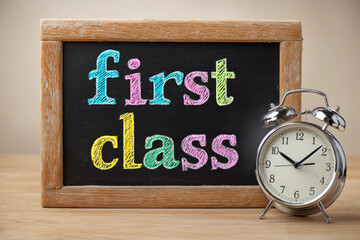 Chalk board with 'First class' sign and an alarm clock on school classroom background. Education, studies and knowledge day concept. Subject shot for poster, demonstration