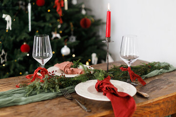 Merry Christmas! Stylish table setting for christmas family dinner. Modern plate and cutlery with napkin, glassware, green and red ribbons, fir branches and candle on rustic table at xmas tree