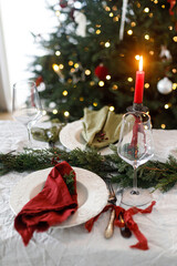 Merry Christmas! Stylish table setting for christmas family dinner. Modern plate and cutlery with napkin, glassware, green and red ribbons, fir branches and candle on rustic table at xmas tree