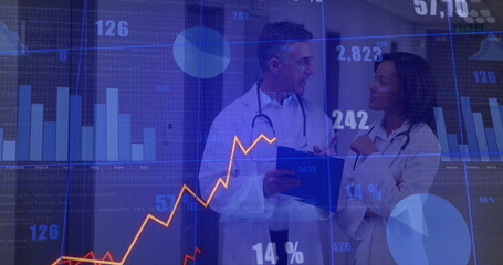 Doctors wearing lab coats and stethoscopes reviewing clipboard in hospital corridor, with charts