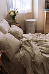 Cozy bedroom scene with oatmeal linen bedding, pillows and a glass vase of fresh flowers on a wicker nightstand, soft natural light.