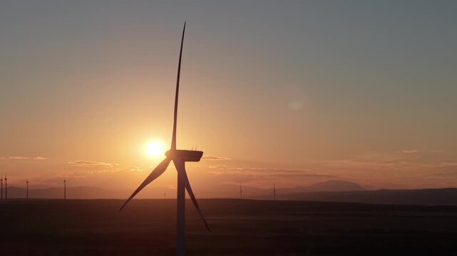 A wind turbine stands tall against the backdrop of a glowing sunset, with mountains and additional turbines visible in the distance. The scene captures the harmony between renewable energy and nature