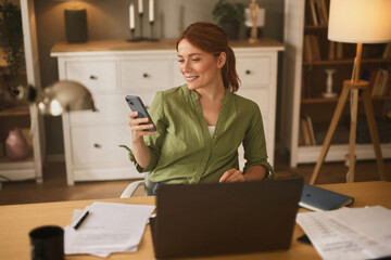 A woman with shoulder-length hair smiles as she looks at her smartphone while seated at a wooden desk in a warmly lit home office filled with books and papers.