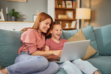 A mother and her daughter share a joyful moment while using a laptop in their inviting living room, filled with a sense of connection and happiness.
