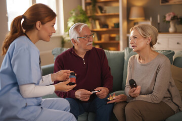 A home health aide is explaining medication dosages to a senior couple at their residence. The nurse is reviewing a pill bottle and blister pack with them as they sit on the sofa in their living room.