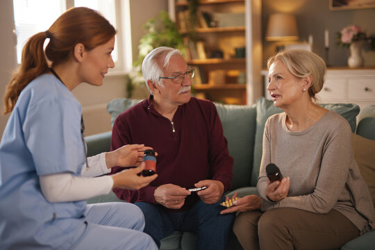 A home health aide is explaining medication dosages to a senior couple at their residence. The nurse is reviewing a pill bottle and blister pack with them as they sit on the sofa in their living room.