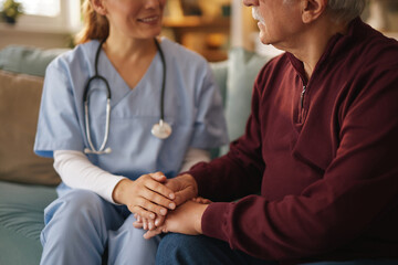 A kind Nurse is holding the hands of a senior man wearing a maroon shirt. They are sitting on a teal sofa during a home care visit in the Living Room of his house during the daytime.