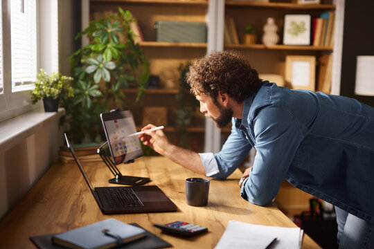 A man is working intently at a home office, using a tablet and stylus while standing by a wooden desk. Natural light pours in from the window, enhancing the workspace ambiance.