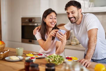 A couple sits at a kitchen table with breakfast dishes, smiling as they interact with a smartphone. They share a joyful moment amid a warm and inviting atmosphere.