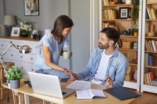 A couple engaged in conversation at a home office, the woman handing a coffee cup to the man, who is reviewing documents on his laptop. Bookshelves and plants add a warm touch. - Powered by Adobe
