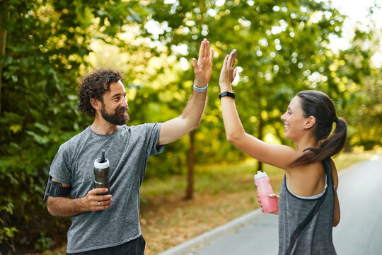 Two friends jog together in a lush park, celebrating their active lifestyle with a cheerful high five and water bottles in hand, embodying fitness and friendship during the morning.