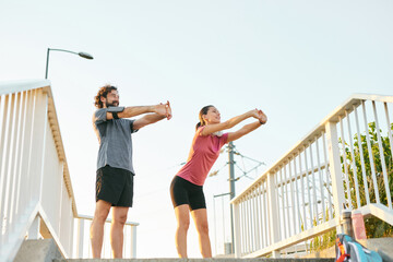 Two people are engaged in stretching exercises on a pedestrian bridge during early morning. Their...