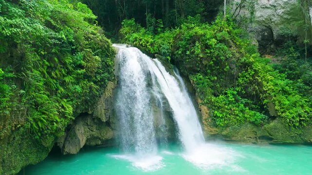 A waterfall flows into a turquoise pool, surrounded by dense tropical jungle and rocky cliffs at Kawasan Falls, Cebu, Philippines.