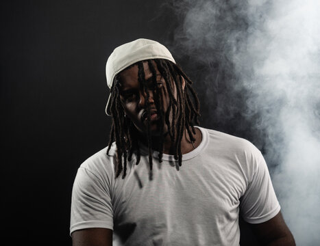 man with dreadlocks wearing a hat and white shirt posing for the camera in a studio against a smoky background.