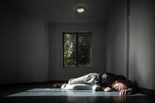The High and The Low. A man lying on the floor in a dimly lit hallway. The image captures the loneliness, isolation, and desperation of drug addiction and homelessness.