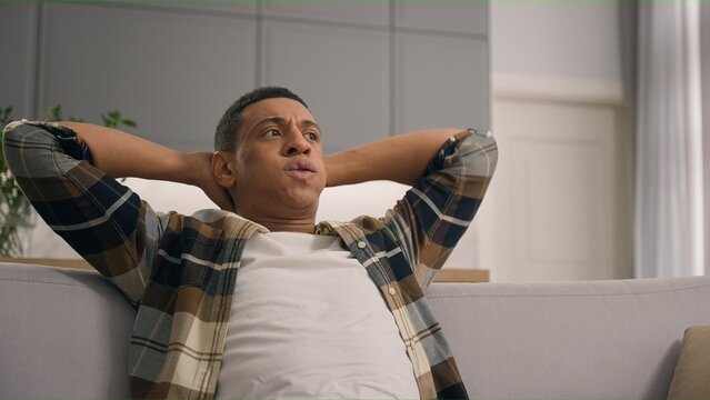 Calm serious breathing African American man relaxed guy ethnic male relaxing at home sofa comfortable couch leaning back hands behind head breath thinking pondering resting in living room apartment