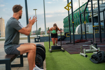 Obraz premium Sporty young woman pushes a weighted sled on artificial turf while a man cheers her on at an outdoor gym.