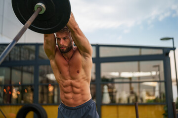 Caucasian fit man performing a barbell shoulder press exercise outdoors. He is shirtless and focused, with a cityscape in the background.