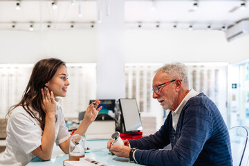 Young and beautiful audiologist showing hearing aid device to senior man in hearing center, explaining how it works and helping him choose the right one for his needs