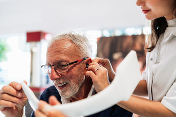 Audiologist is helping a senior man put on a hearing aid. While he looks in a mirror to see the process in a modern clinic setting. Showcasing the importance of hearing healthcare for the elderly