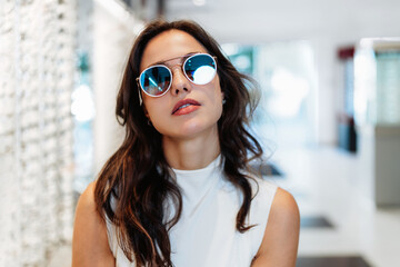 Young woman trying on blue sunglasses in an optical store, touching her face and making a decision...