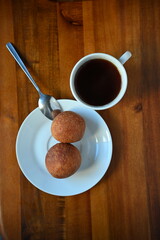 A top view photograph of two homemade doughnuts and a cup of black coffee on a white plate with a spoon set on a rustic wooden table. The image captures a simple, cosy breakfast or snack.