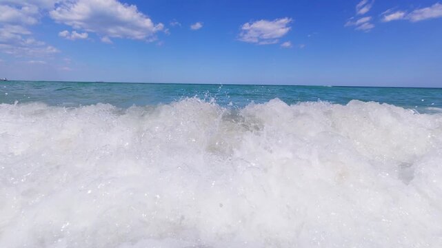 Slow motion POV shot of powerful ocean wave crashing near shore, white foam and clear turquoise water under blue sky with scattered clouds