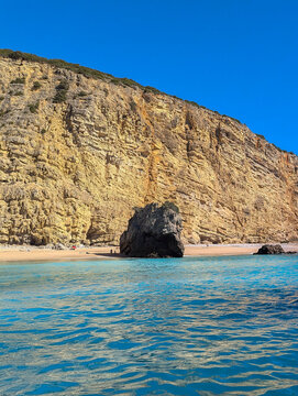 Black rock on sandy beach in front of cliff with turquoise blue sea water in Sesimbra, Portugal