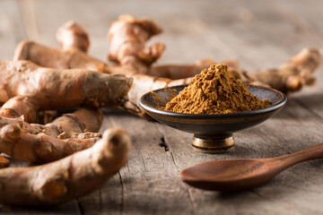 Ground turmeric powder in a bowl with raw turmeric root on wood