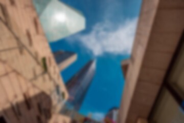 An abstract, bokeh-filled view of a bright blue sky with white clouds, framed by buildings and looking upward.