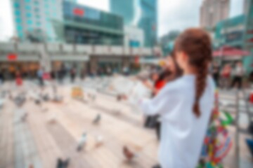 Obraz premium A blurry image of a woman with long brown hair walking in front of a modern shopping mall in a bustling city.