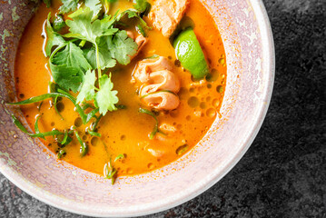 Spicy Thai soup with orange broth, chicken, lime, cilantro, and herbs in a ceramic bowl on dark background. Traditional Asian cuisine, close-up, top view