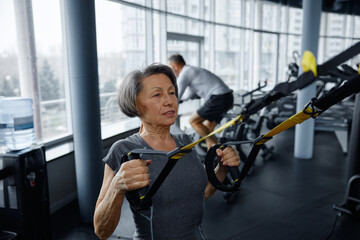 Mature woman is exercising on a suspension trainer in a gym