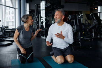 Elderly man and woman sit on yoga mats in a gym talking