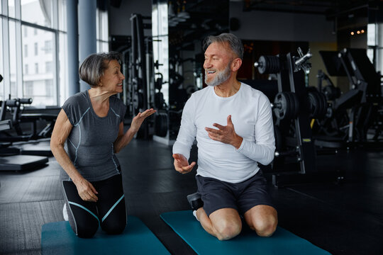 Elderly man and woman sit on yoga mats in a gym talking - Powered by Adobe