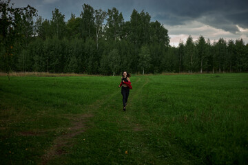 girl dressed in black pants and a red top runs joyfully across a lush green field. sky is overcast, hinting at impending rain, and trees line the edge of the meadow